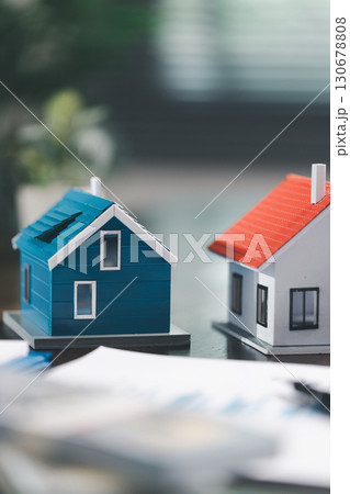 Real estate agent and customer are discussing contract to buy house, with house model in background symbolizing real estate, insurance, and loan arrangements involved in process. Real estate agent and customer are discussing contract to buy house, with house model in background symbolizing real estate, insurance, and loan arrangements involved in process. 130678808