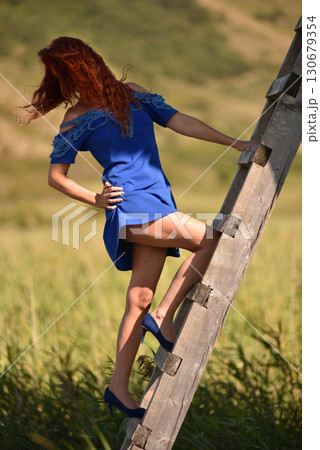Red haired woman in vibrant blue dress and high heels ascends a rustic wooden ladder in the outdoors Red haired woman in vibrant blue dress and high heels ascends a rustic wooden ladder in the outdoors 130679354