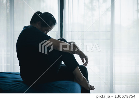 A stressed woman sitting alone on bed in dark room, holding her head down, symbolizing depression, anxiety, hopelessness, loneliness, mental health struggle, burnout and emotional pain. 130679754