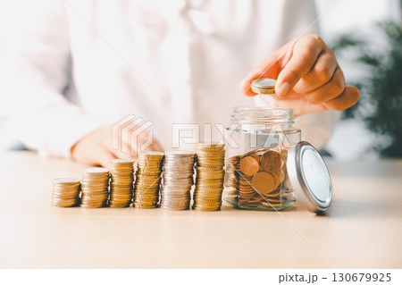 Hand places coin into saving jar, representing how smart finance, investment, banking practices turn money, cash, currency into financial wealth drives business growth and strengthens the economy. Hand places coin into saving jar, representing how smart finance, investment, banking practices turn money, cash, currency into financial wealth drives business growth and strengthens the economy. 130679925