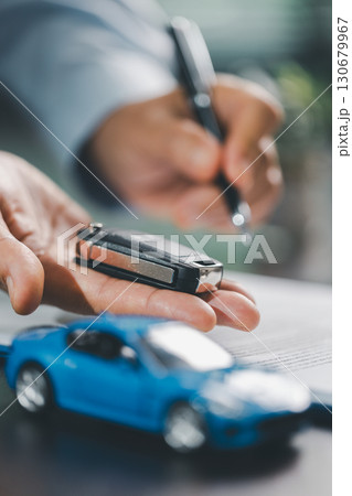 Business office, woman employee finalizes deal with client, handing over car key after signing contract and reviewing insurance and loan documents for sale or rent of vehicle at table. Business office, woman employee finalizes deal with client, handing over car key after signing contract and reviewing insurance and loan documents for sale or rent of vehicle at table. 130679967