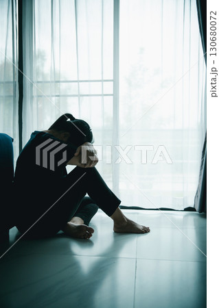 A lonely woman sits on the floor by window with hand on head in dark room. Concept of depression, sadness, anxiety, stress, mental health problem, loneliness and emotional struggle in life. A lonely woman sits on the floor by window with hand on head in dark room. Concept of depression, sadness, anxiety, stress, mental health problem, loneliness and emotional struggle in life. 130680072