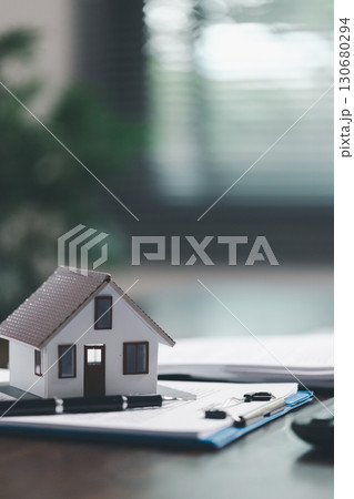 Real estate agent and customer are discussing contract to buy house, with house model in background symbolizing real estate, insurance, and loan arrangements involved in process. 130680294