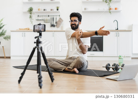Young adult man performing fitness arm stretches while recording video blog in home setting surrounded by exercise equipment showing lifestyle and health routine. 130680331