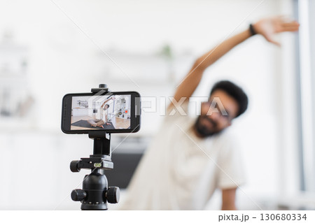 Young adult man records fitness tutorial at home using smartphone camera, focusing on arm stretches. Promotes health, fitness, and active lifestyle through shared video content on personal platforms 130680334