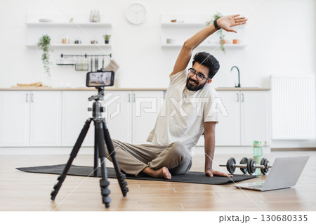 Young adult man stretching with arms raised, recording fitness blog in home environment. Shot features home decor, fitness equipment, water bottle, and a motivational approach to healthy living 130680335