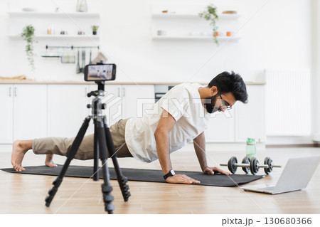 Young bearded man wearing casual clothes exercising on yoga mat at home recording fitness video while performing push-ups. Camera and laptop nearby show dedication to health and social media content. 130680366
