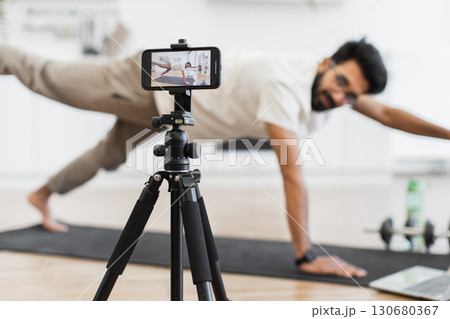 Man performing yoga pose while recording content for a fitness blog using smartphone setup on tripod. Promotes exercise, mindfulness, and home health routines for an active lifestyle improvement. 130680367