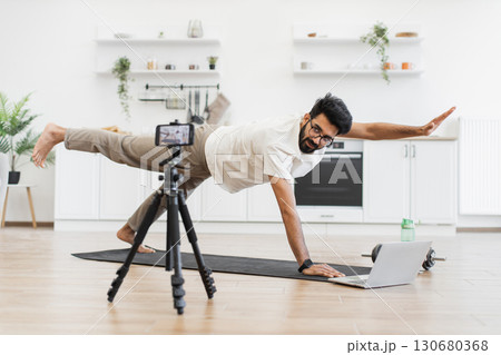 Man showcasing yoga pose during online blogging session in home kitchen, maintaining balance. Middle-aged male focused on fitness, engaging audience virtually emphasizing healthy lifestyle 130680368