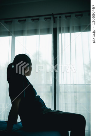 A woman sitting alone on bed facing window in dark room, symbolizing depression, loneliness, anxiety, sadness, mental health struggle, insomnia and emotional stress in life challenges. 130680467