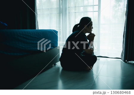 A woman sitting on the floor near window with hands clasped in thoughtful pose. Concept of depression, loneliness, anxiety, prayer, hope, mental health, emotional struggle and self-reflection. 130680468