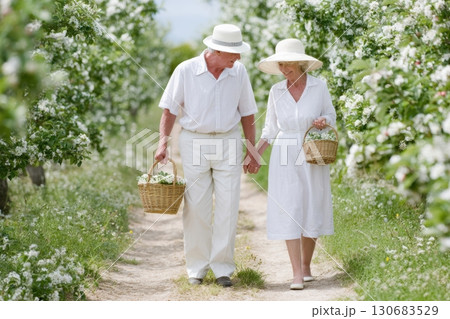 Senior couple walking through a blooming orchard holding hands and carrying baskets of white flowers, dressed in white clothes and sun hats on a sunny day 130683529