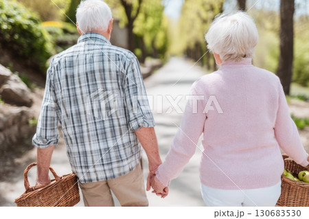 An elderly couple walks hand-in-hand down a peaceful park path on a sunny day, each carrying a wicker basket. A serene and intimate moment of companionship An elderly couple walks hand-in-hand down a peaceful park path on a sunny day, each carrying a wicker basket. A serene and intimate moment of companionship 130683530