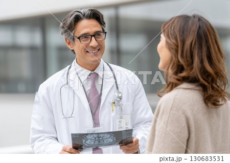 A smiling male doctor shows a brain scan to a female patient during a consultation outside a medical facility. Friendly and professional healthcare interaction A smiling male doctor shows a brain scan to a female patient during a consultation outside a medical facility. Friendly and professional healthcare interaction 130683531