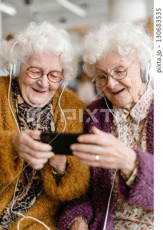Two elderly women with white curly hair, wearing headphones and colorful sweaters, smiling and watching something on a smartphone together 130683535