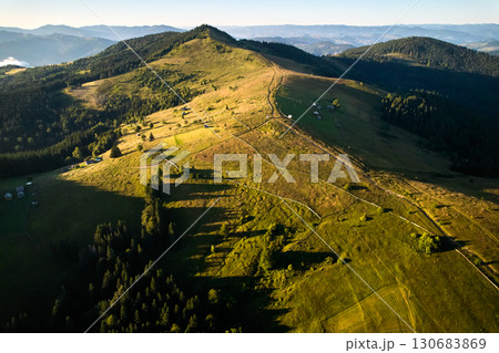 Aerial view of verdant mountain landscape bathed in morning light. Rolling hills with scattered farmhouses interspersed with patches of forest. Sunlight creates long shadows. Aerial view of verdant mountain landscape bathed in morning light. Rolling hills with scattered farmhouses interspersed with patches of forest. Sunlight creates long shadows. 130683869