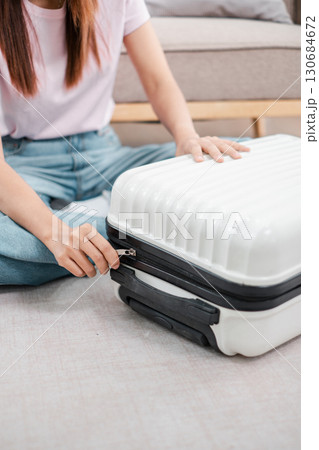 A woman in casual clothing zips a white suitcase on the floor, preparing for travel. 130684672