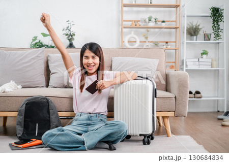 Joyful young woman with passport and suitcase, ready for travel adventure, sitting in a stylish living room. 130684834