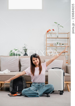 Smiling woman sitting on floor with suitcase and backpack, ready for travel. Modern living room with plants and decor. 130684835