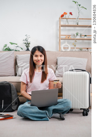 Woman sitting on floor with laptop and credit card, ready for travel. Cozy home setting with plants and luggage. Woman sitting on floor with laptop and credit card, ready for travel. Cozy home setting with plants and luggage. 130684836
