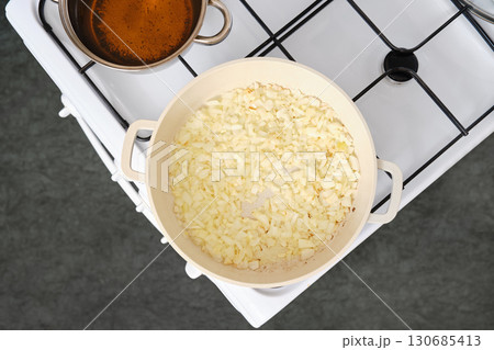Chopped onions cooking in a pan on a stove with simmering pot of broth nearby 130685413