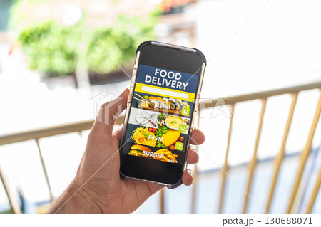 Hand holding smartphone with food delivery app interface on screen, showing sushi, vegan, pasta and burger categories. Hand holding smartphone with food delivery app interface on screen, showing sushi, vegan, pasta and burger categories. 130688071