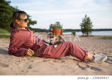 Relaxed woman lounging on sunny beach with uneasy cat curled on lap. Feline travel adaptation 130690917
