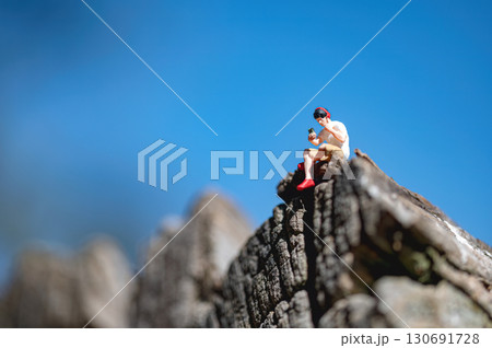 Miniature man sitting on a rock using a smartphone and listening music in headphones with a blue sky background 130691728