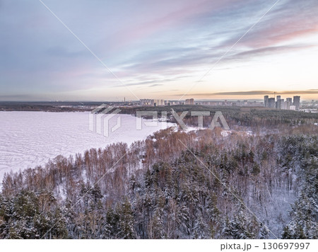 Snow-covered forest on lake shore with the first ice at sunset and the city on horizon, aerial view Snow-covered forest on lake shore with the first ice at sunset and the city on horizon, aerial view 130697997