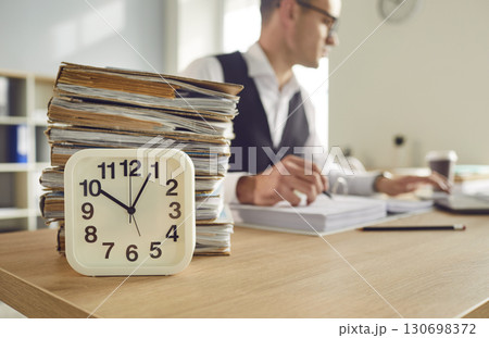Overworked tired man accountant working in office with clock and pile of folders in foreground. Overworked tired man accountant working in office with clock and pile of folders in foreground. 130698372