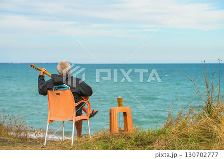 Elderly man playing guitar while sitting on chair near seaside cliff with beer on table. Concept of solitude, coastal peace, creative escape, musical freedom Elderly man playing guitar while sitting on chair near seaside cliff with beer on table. Concept of solitude, coastal peace, creative escape, musical freedom 130702777