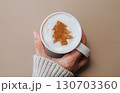 Hand in cozy sweater holding coffee mug with Christmas tree latte art on milk foam, festive holiday mood 130703360