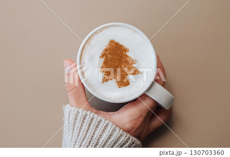 Hand in cozy sweater holding coffee mug with Christmas tree latte art on milk foam, festive holiday mood 130703360