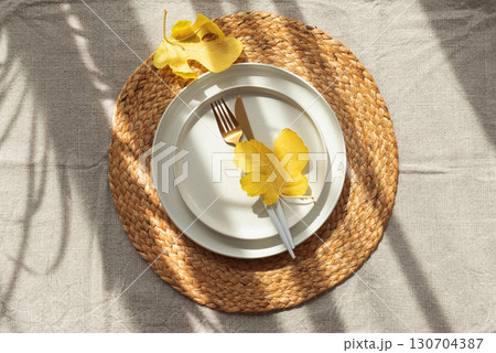 Table Setting with Yellow Leaves on White Plate and Woven Placemat under Sun Light Table Setting with Yellow Leaves on White Plate and Woven Placemat under Sun Light 130704387