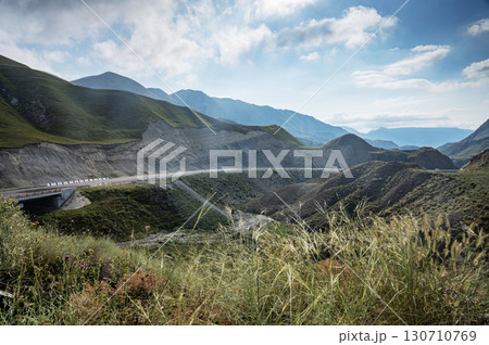 Mountainous terrain with deep valleys, green slopes and winding road under cloudy blue sky. 130710769