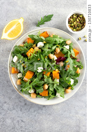 Healthy arugula salad with roasted pumpkin, chickpeas, feta cheese, pistachios on gray background. Healthy arugula salad with roasted pumpkin, chickpeas, feta cheese, pistachios on gray background. 130710833