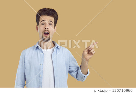 Man with funny shocked facial expression pointing finger to right side isolated on beige background. Man with funny shocked facial expression pointing finger to right side isolated on beige background. 130710960