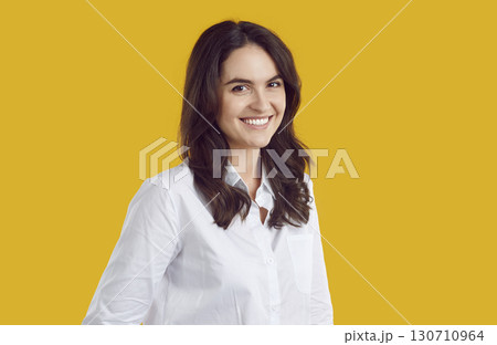 Smiling shy brunette woman in white shirt is posing on yellow background looking at camera. Smiling shy brunette woman in white shirt is posing on yellow background looking at camera. 130710964