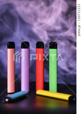 A row of colorful disposable vapes on a black surface against a smoky background. 130712151