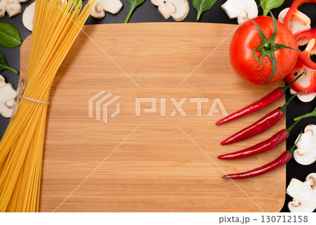 A cutting board, raw spaghetti, fresh tomato, chili peppers, and mushrooms, framed on a dark background. A cutting board, raw spaghetti, fresh tomato, chili peppers, and mushrooms, framed on a dark background. 130712158