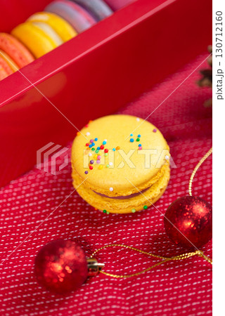 A bright yellow macaron with sprinkles, alongside red Christmas ornaments and a gift box, on a festive red background 130712160