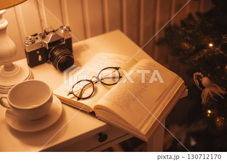 A cozy nightstand with an open book, reading glasses, a vintage camera, and a cup, all bathed in warm, soft light. 130712170