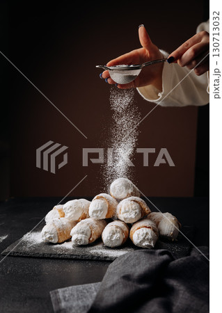 Fresh cream horns being dusted with powdered sugar on dark background, winter dessert vibes Fresh cream horns being dusted with powdered sugar on dark background, winter dessert vibes 130713032