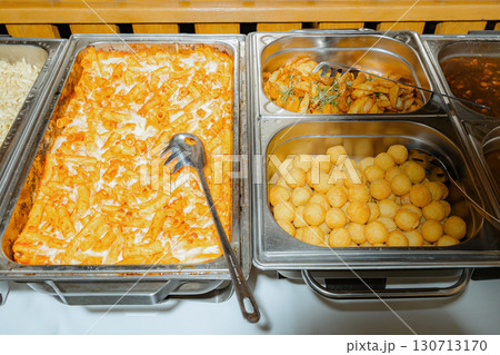 Buffet setup featuring baked pasta, crispy potatoes, and fried snacks in serving trays. Perfect for events, displaying a tempting variety of dishes for guests to enjoy 130713170