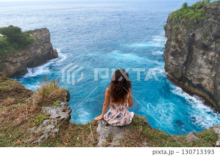 A beautiful woman in a pink dress sits on a cliff above the ocean on the island of Nusa Penida. Devil's Billabong an incredibly wonderful lagoon with splashes from the waves. 130713393