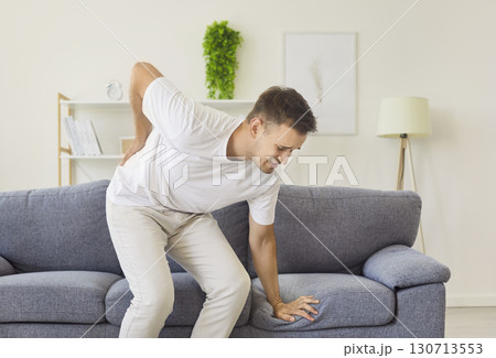 Man suffering sharp lower back pain while standing up from couch at home Man suffering sharp lower back pain while standing up from couch at home 130713553