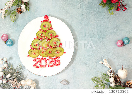 Winter background for greeting card. Edible Christmas tree made of kiwi and pomegranate on the festive table, idea for food design and decoration of New Year's dishes for children. 130713562