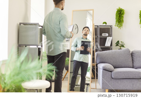 Young man looking at the mirror preparing for work standing at home and holding documents in hands Young man looking at the mirror preparing for work standing at home and holding documents in hands 130715919