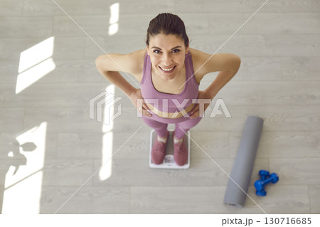 Happy fit young woman does sports exercises and weighs herself on scales at home Happy fit young woman does sports exercises and weighs herself on scales at home 130716685