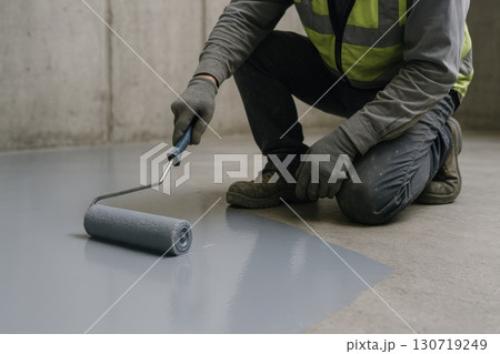 Worker applying protective floor coating with paint roller on concrete surface, wearing gloves and safety vest during industrial renovation and construction work 130719249
